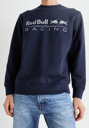 Osoba nosząca granatową bluzę z białym logo Red Bull Racing i jasnoniebieskie dżinsy na jednolitym tle.