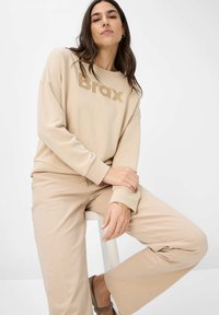 Beige sweatshirt med "Brax" logo i en dæmpet tone, med en afslappet pasform og brede ærmer. Parret med matchende beige bukser og sandaler.