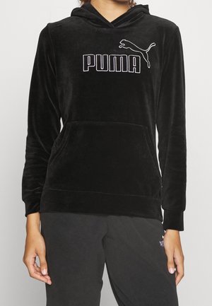 Osoba ubrana w czarną welurową bluzę z kapturem z białym logo Puma na piersi oraz pasujące czarne spodnie, stojąca na jednolitym tle.