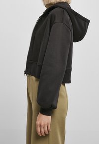 Black cropped hoodie with a zipper, large hood, and ribbed cuffs. Made of soft fabric; paired with light olive green pants.