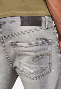 Utblekta grå denimjeans med slitna detaljer, med en bakficka som har metallnitar och en bälteetikett i svart läder.
