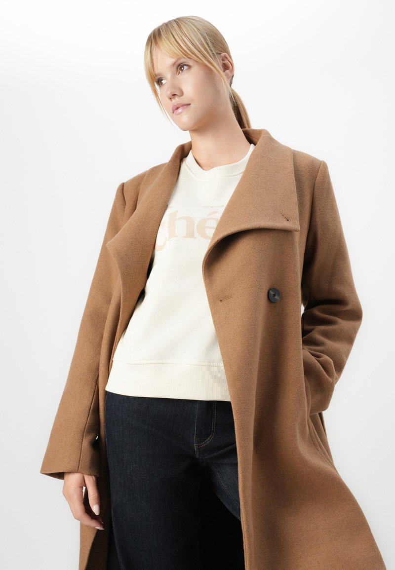 Cappotto di lana marrone con colletto grande, chiusura a un bottone e tasche laterali indossato sopra un maglione crema e jeans scuri.