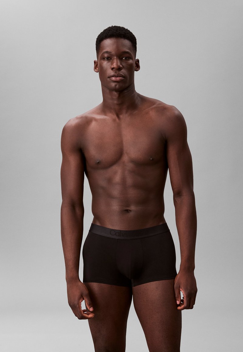 Czarne bokserki z miękkim paskiem w talii z logo Calvin Klein, model pozuje na neutralnym tle, skupiając się na dopasowaniu i materiale.