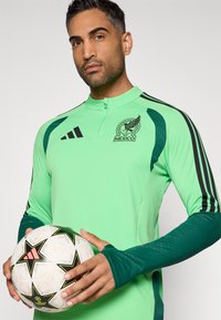 Πράσινη ποδοσφαιρική φανέλα με μακριά μανίκια και κεντημένο έμβλημα του Μεξικού, μαύρες ρίγες και αντίθετους σκούρους πράσινους τόνους. Κρατάει μια πολύχρωμη ποδοσφαιρική μπάλα.