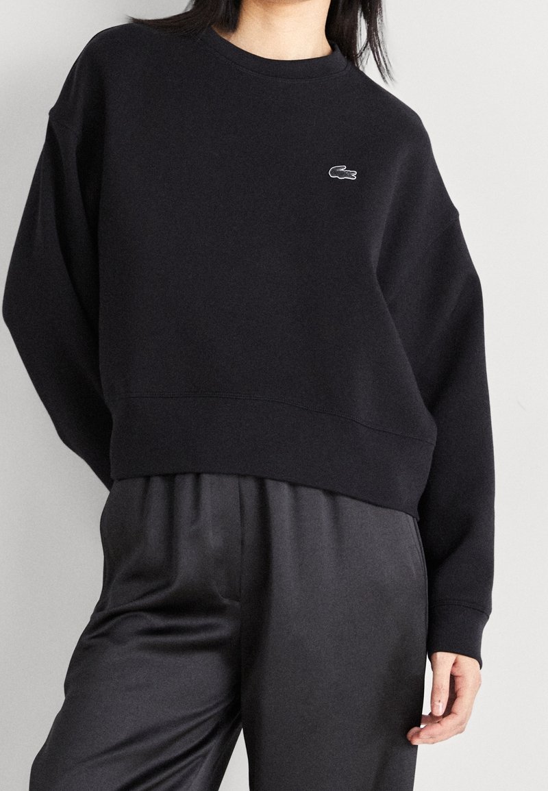 Personne portant un sweat-shirt noir à manches longues avec un petit logo crocodile blanc sur la poitrine et un pantalon noir ample sur un fond uni.