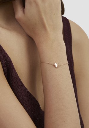 Armbånd i rosaguld med en lille hjertevedhæng på en delikat kæde, båret på et bart håndled mod en mørk stofbaggrund.