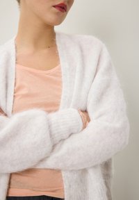 Valkoinen, pörröinen kardigan, jossa on avonainen etuosa ja pitkät hihat, kerroksittain vaaleanpunaisen V-aukkoshirtin päällä. Pehmeä rakenne, rento istuvuus.