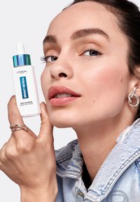 Bouteille de sérum Bright Reveal, blanche avec un capuchon teal. Le modèle la tient près de son visage, mettant en valeur une peau lisse et des bijoux en argent avec une veste en denim.