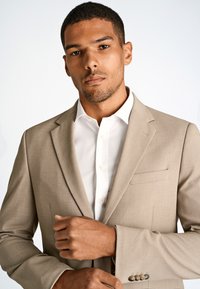 Giacca beige con finitura testurizzata, rever a punta e una tasca sul petto. Camicia bianca con chiusura a bottoni sotto.