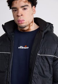 Chaqueta de plumas negra con capucha, presenta ribetes blancos. Lleva puesta una sudadera azul marino con el logotipo de "ellesse" en blanco y acentos rojos.