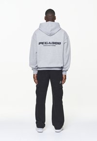 Grå hoodie med texten "PEGADOR" och "CERTIFIED AUTHENTIC STREETWEAR" nedanför, tillsammans med svarta cargo byxor och vita sneakers.