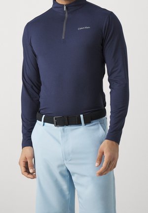 Hombre con camiseta de manga larga azul marino con cierre de un cuarto de cremallera y logo de Calvin Klein, y pantalones azul claro con cinturón negro tejido.