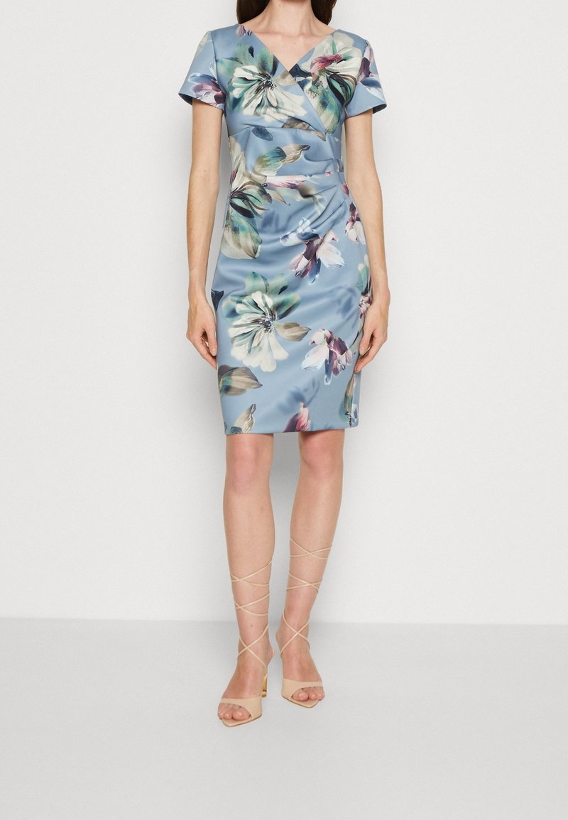 Femme portant une robe bleue cintrée au genou avec un grand imprimé floral, manches courtes, et des sandales beiges à talons hauts et à brides.