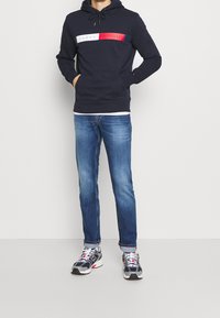 Muž v námořnickém mikině s logem Tommy Hilfiger, modrých džínách a bílých a černých teniskách New Balance, stojící s rukama v kapsách.