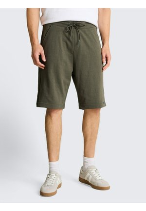 Mann trägt olivgrüne Shorts mit Kordelzug, weiße Socken und beige Sneaker, steht vor einem schlichten hellen Hintergrund.