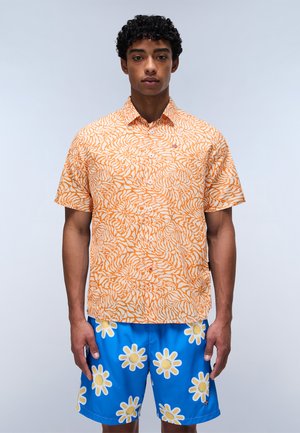 Camisa de manga corta en color naranja con un patrón abstracto en blanco y ondulado. Combinada con pantalones cortos azules con estampado de margaritas amarillas.
