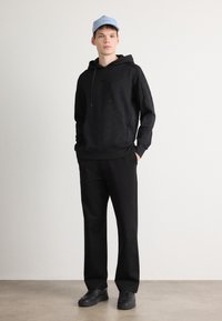 Μαύρα φούτερ με κορδόνια, μπροστινή τσέπη, συνδυασμένα με μαύρο παντελόνι και μαύρα αθλητικά παπούτσια. Ανοιχτό μπλε καπέλο με λεπτομέρειες κειμένου. Μινιμαλιστικός σχεδιασμός.