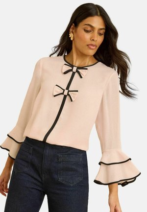 Blus i mjukt rosa tyg, med svart kant, två dekorativa rosetter och utsvängda ärmar med lager av volanger. Bärs med mörka jeans.