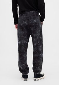 Czarne spodnie dresowe tie-dye z elastycznym pasem i ściągaczami u dołu. Posiadają tylną kieszeń i miękki, teksturowany materiał. Zestawione z czarnymi sneakersami.