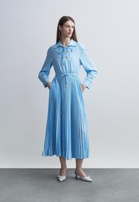 Femme debout les mains dans les poches, portant une robe midi à manches longues rayée bleu clair et des talons blancs à bouts pointus.