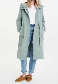 Light green, long raincoat with a hood, adjustable drawstrings, front pockets, and snap-button closure. Paired with blue jeans and brown loafers.