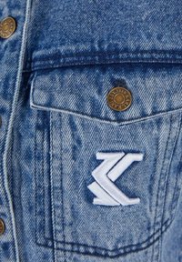Primer plano de un bolsillo de chaqueta de mezclilla azul con un botón de latón etiquetado "KAREY" y un logo bordado en blanco con la "K" en la tela.
