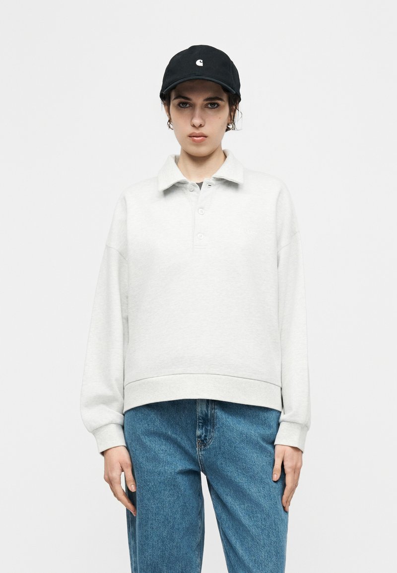 Persona joven con gorra negra, polo de manga larga gris claro y vaqueros azules, de pie contra un fondo blanco liso.