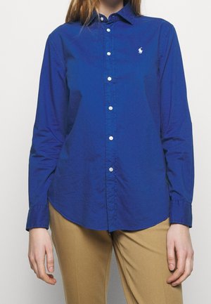 Persona vistiendo una camisa de manga larga azul real abotonada con un logo blanco en el pecho y pantalones marrón claro, de pie con los brazos relajados.