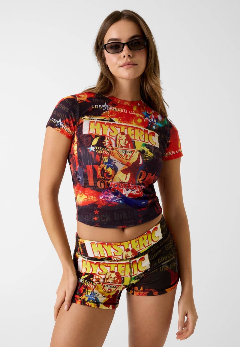 Jeune femme portant des lunettes de soleil noires, un crop top graphique multicolore et un short assorti avec les inscriptions « HYSTERIC » et « GUESS ».