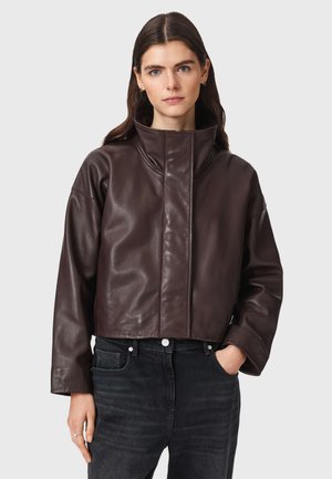 Frau mit langen dunklen Haaren trägt eine dunkelbraune Lederjacke mit hohem Kragen und schwarze Jeans, steht vor einem schlichten Hintergrund.