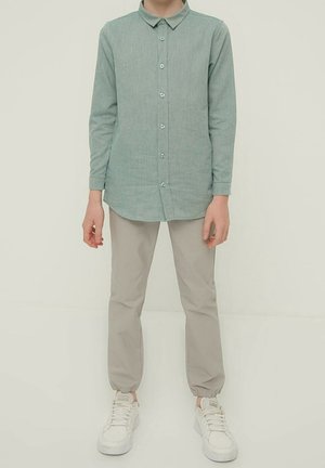 Person iført en grøn langærmet skjorte med knapper, beige bukser med elastisk manchet og hvide sneakers, der står mod en ensfarvet baggrund.