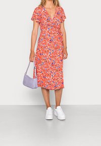 Robe portefeuille rouge à fleurs avec manches volantées, agrémentée d'accents bleus et blancs. Associée à un sac à main lavande et des baskets blanches.