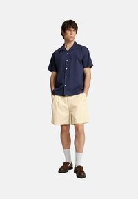 Camicia a maniche corte della Marina, pantaloni di blu avorio, calzini bianchi, scarpe marroni. Il modello è in piedi con le mani in tasca su uno sfondo semplice.