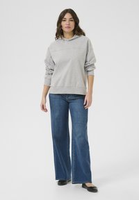 Sudadera gris con capucha y mangas largas, combinada con jeans de mezclilla azul de pierna ancha. El atuendo se complementa con zapatos planos negros.