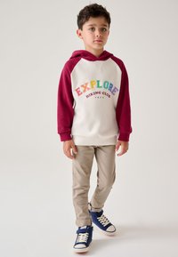 Kapučiniškas džemperis kreminės ir bordo spalvų su daugiaraščių raidžių „EXPLORE“ tekstu; derinamas su smėlio spalvos kelnėmis ir mėlynomis sportinėmis batelėmis su baltais padais.