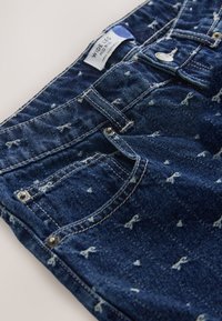 Jeansy z szerokimi nogawkami w ciemnym niebieskim kolorze z efektem sprania. Posiadają srebrne okucia, liczne kieszenie oraz metkę na pasku w talii.