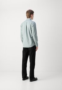 Lichtblauw overhemd met lange mouwen, met een rugplooidetail, gecombineerd met zwarte broeken en zwarte schoenen, staande tegen een witte achtergrond.