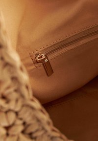 Beżowa tkanina wewnątrz torby z kieszenią zapinaną na zamek błyskawiczny w złotym kolorze. Miękka tekstura i przeszywane krawędzie. Widoczny wzór tkany.