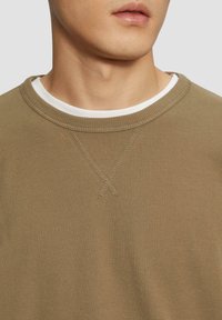 Khakifarbener Pullover mit strukturiertem, geripptem Finish. Verfügt über einen weiten Ausschnitt und ein Kreuzstichdetail am Kragen. Über einem weißen Hemd getragen.