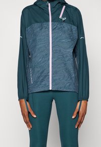 Teal udendørs vindjakke med et mønstret brystpanel, kontrastfarvet lyserød lynlås, justerbare manchetter og hætte, lavet af letvægtsmateriale.