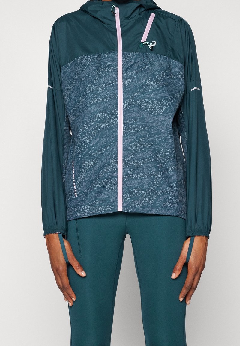 Teal udendørs vindjakke med et mønstret brystpanel, kontrastfarvet lyserød lynlås, justerbare manchetter og hætte, lavet af letvægtsmateriale.