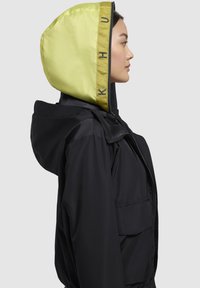 Veste noire avec une capuche jaune vif présentant un tissu texturé et des accents de logo. La vue latérale met en avant une fermeture éclair et des poches spacieuses.