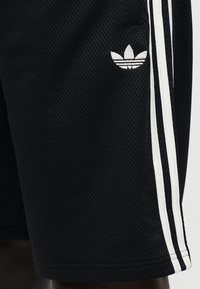 Juodi sportiniai šortai iš tinklelio, turintys baltą trijų juostelių dizainą ir mažą baltą Adidas logotipą šone. Tekstūruota medžiaga, laisvas fasonas.