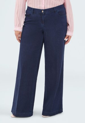 Frau trägt dunkelblaue Jeans mit weitem Bein und ein pink gestreiftes Hemd mit Knöpfen, steht mit entspannten Händen an den Seiten.