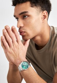 Stainless steel watch with a green dial and silver accents. Features chronograph sub-dials and a textured bezel, worn on a wrist.