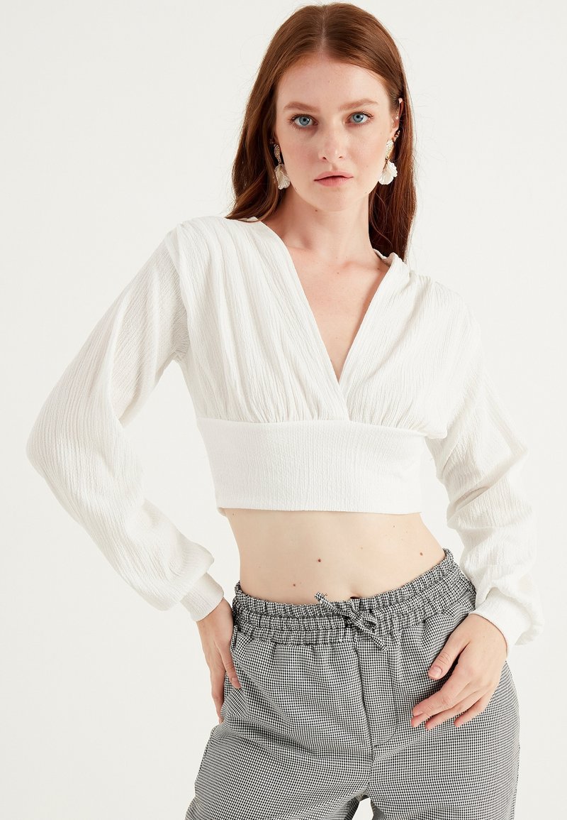 Blusa bianca corta con maniche lunghe, tessuto testurizzato, scollatura a V profonda, abbinata a pantaloni a quadri a vita alta con cintura elastica.