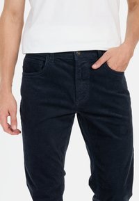Donkerblauwe corduroy broek met een gestructureerd oppervlak, voorzien van steekzakken en een knoopsluiting, gefotografeerd met een hand in een zak.
