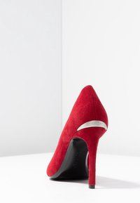 Escarpin en daim rouge avec un bout pointu et un talon aiguille élégant. Présente un accent argenté au niveau du talon et une surface texturée.