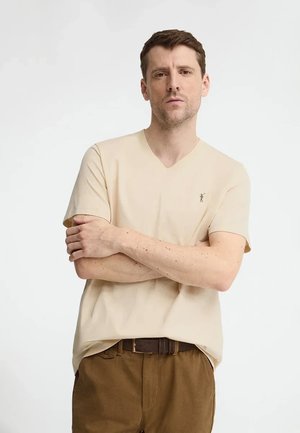 Man met kort bruin haar, gekleed in een beige V-hals T-shirt en bruine broek, staat met gekruiste armen tegen een effen achtergrond.