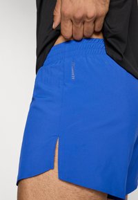 Μπλε αθλητικό σορτς με υφή σαν δικτυωτό πλέγμα, με ελαστική μέση με κορδόνι και πλάγιες σκισμές για ελευθερία κινήσεων. Λογότυπο κοντά στη μέση.